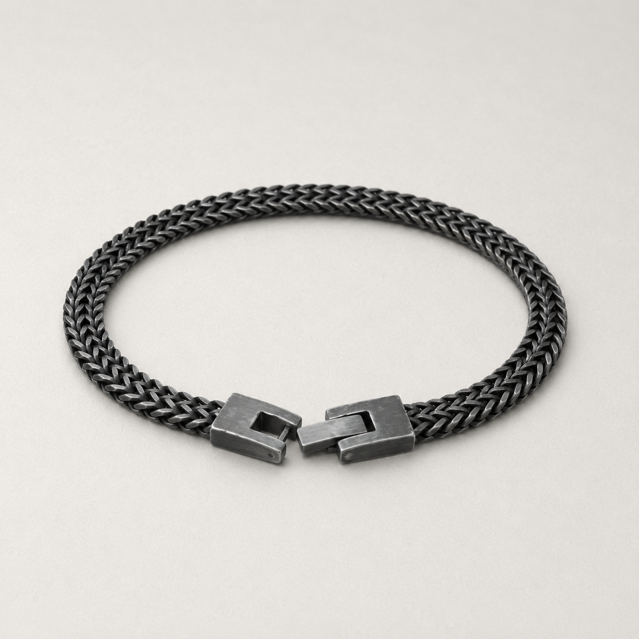 A dark gunmetal braided chain bracelet with a matching geometric slide clasp, displayed on a light gray background.
